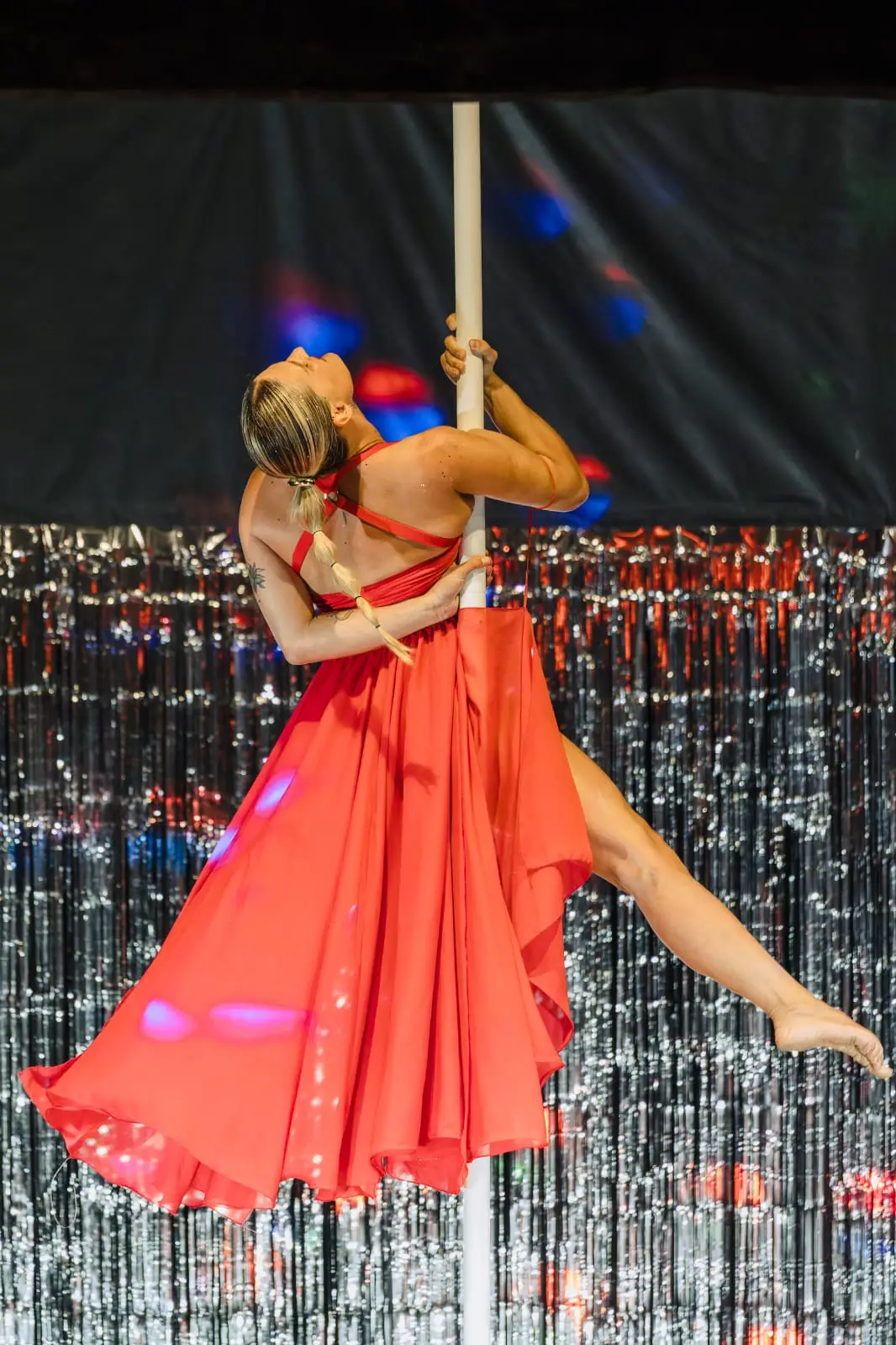 Danseuse en robe rouge fluide accrochée au pôle, tête renversée, sur la scène d’un spectacle de pole dance