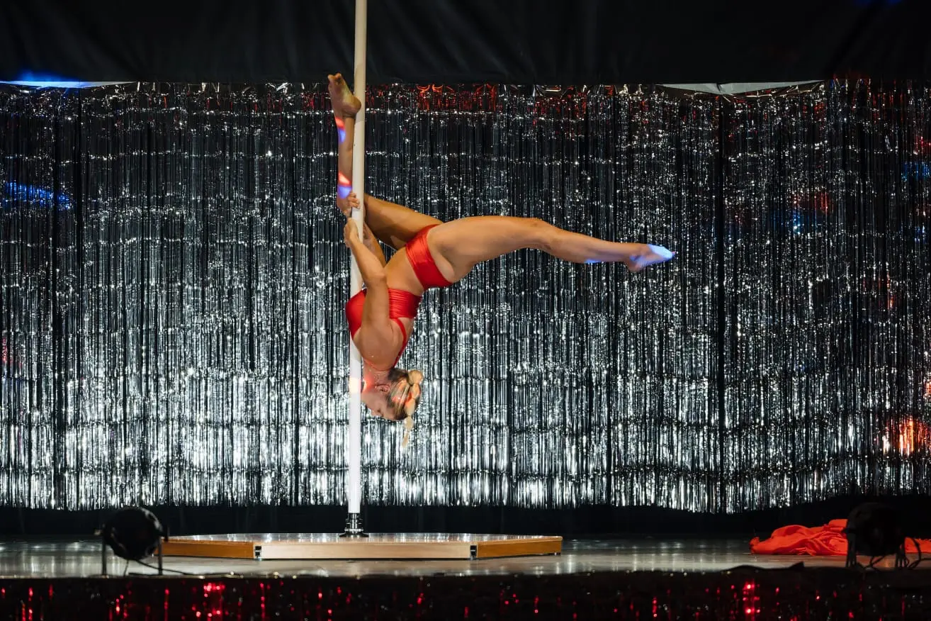 Danseuse en tenue rouge suspendue à l’horizontale le long du pôle, devant un rideau argenté