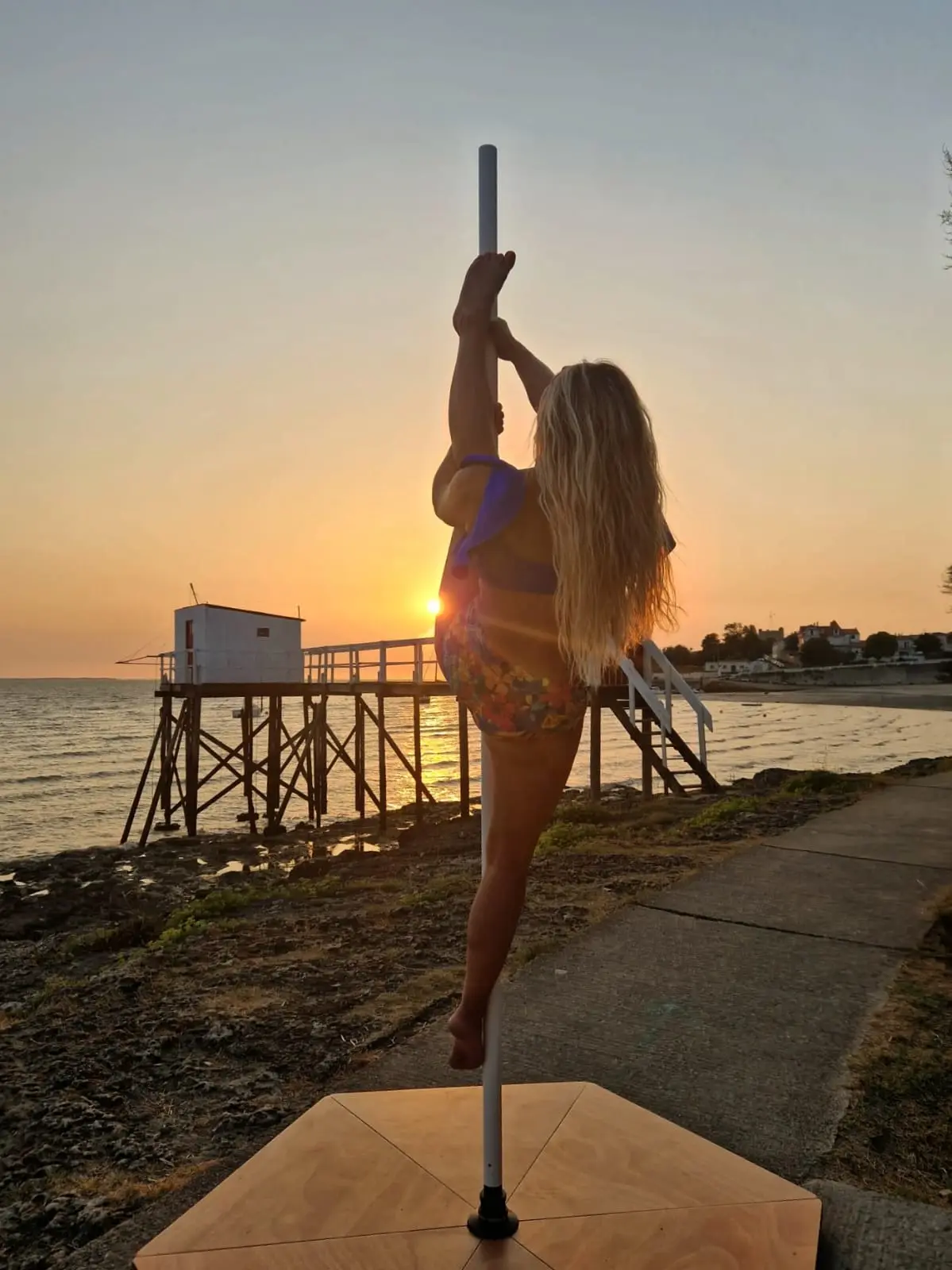 Danseuse en grand écart debout, une main au pôle, face au soleil couchant en bord de mer