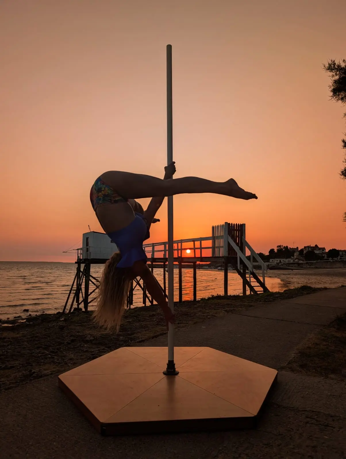 Danseuse en figure inversée sur un pôle, jambes tendues à l’horizontale devant un coucher de soleil sur la mer