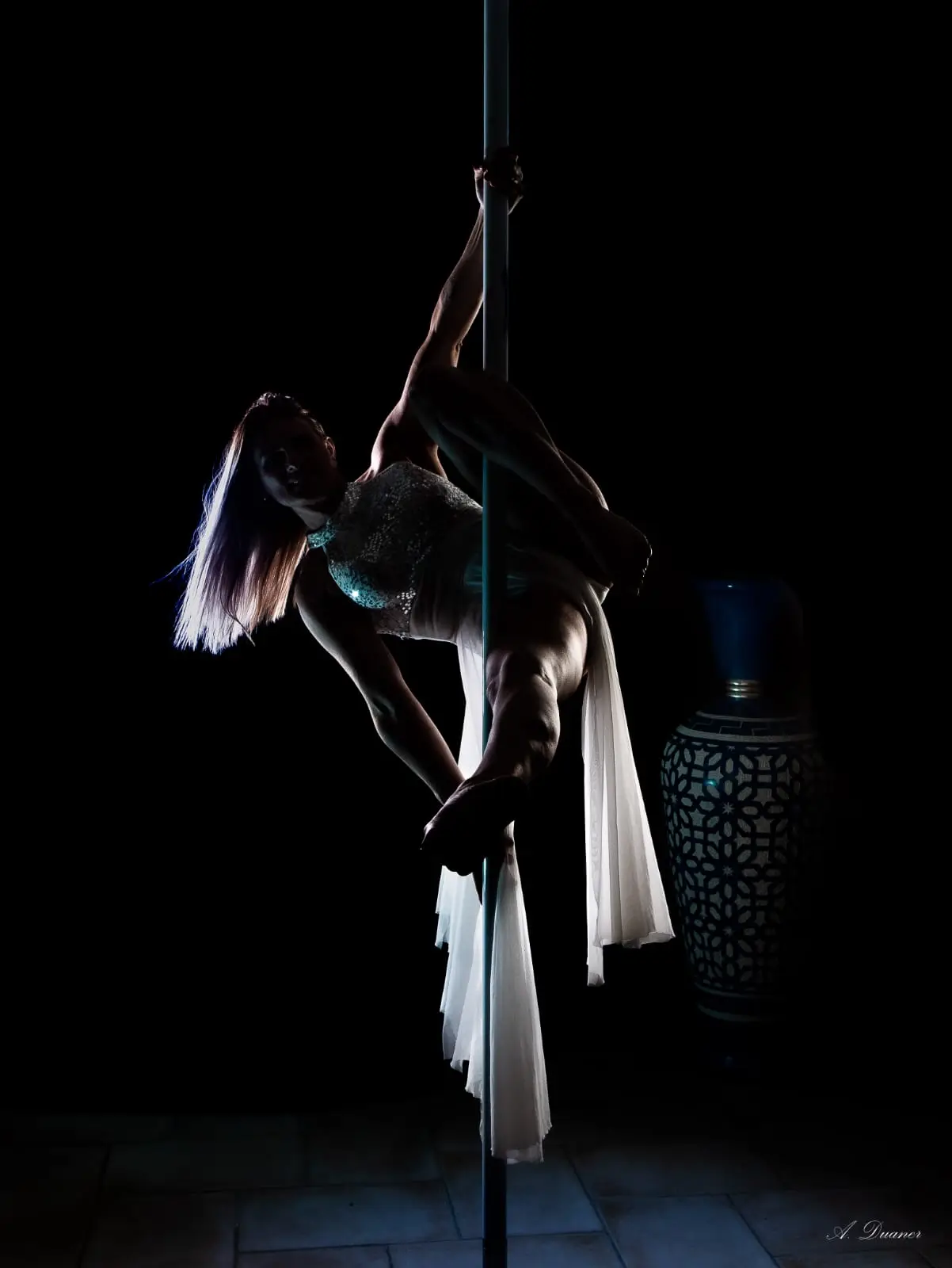 Danseuse en robe blanche suspendue au pôle, éclairée en clair-obscur sur fond noir
