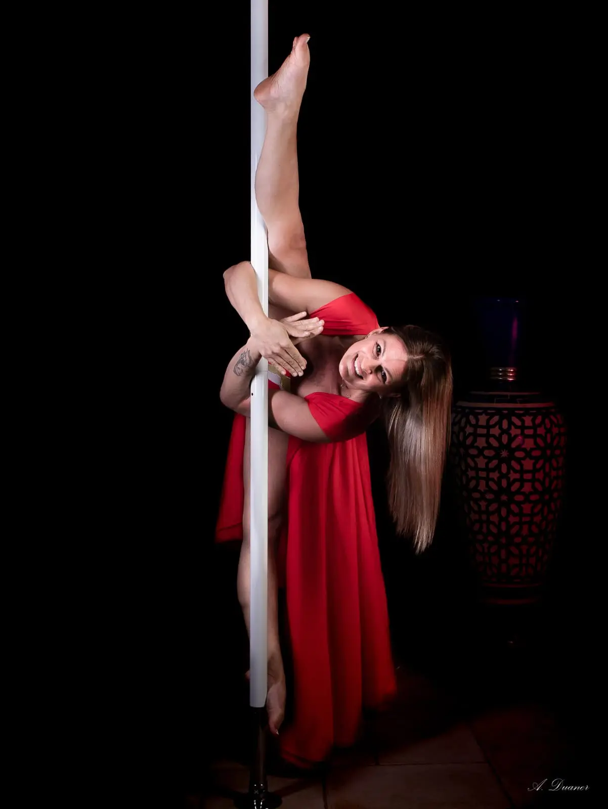 Danseuse en robe rouge au pôle, dans une pièce chaleureuse où l’on distingue une lanterne ajourée et un sol carrelé